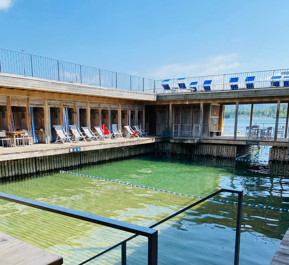 Seebad Lucerne, a popular lakeside swimming area featuring a wooden deck built over Lake Lucerne. Visitors lounge and swim in the designated pools within the lake, surrounded by the structure’s minimalist architecture and open-air design. 