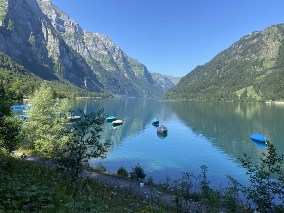 A clear day view of Lake Klöntal, a serene mountain lake in Switzerland, nestled in the Glarus region with towering mountains on both sides reflected in the crystal-clear waters.