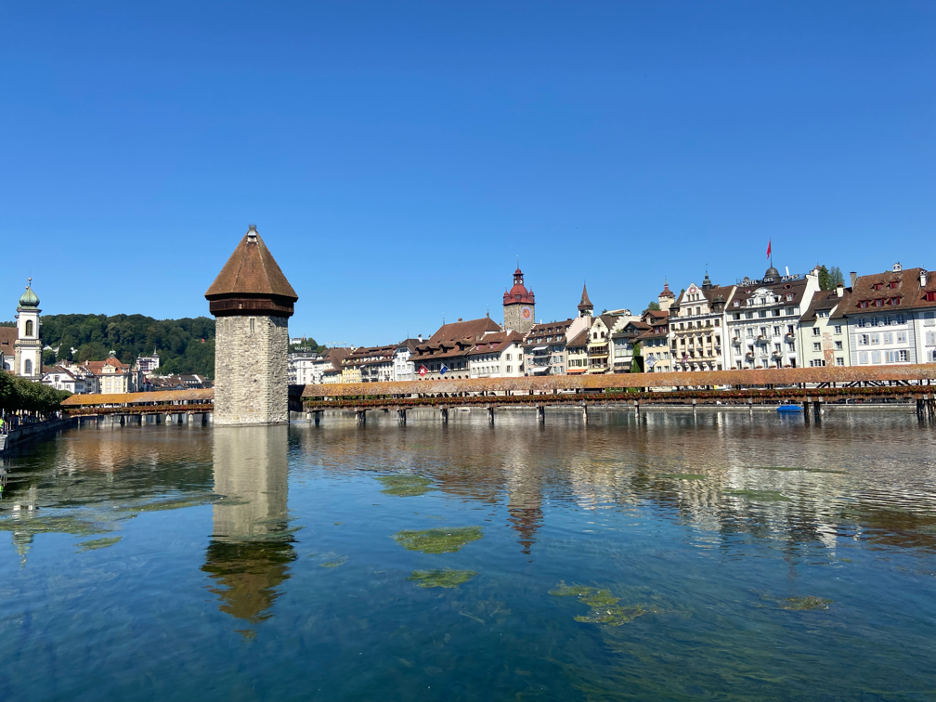 A scenic view showcasing some of the best things to do in Lucerne, Switzerland, on a clear day, featuring the historic Chapel Bridge and its iconic Water Tower. The wooden bridge stretches diagonally across the tranquil Reuss River, adorned with vibrant flower boxes along the railings. The octagonal Water Tower stands nearby, its reflection visible in the calm water, with traditional Swiss architecture as a picturesque backdrop.