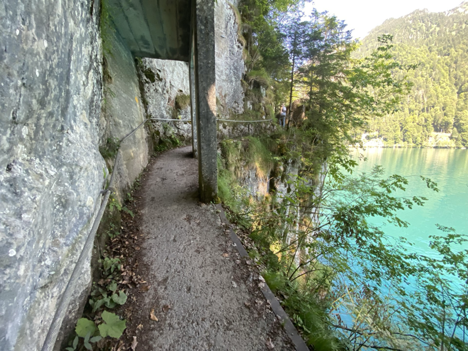 A narrow path winding along a rocky cliffside beside the tranquil waters of Lake Klöntal, a picturesque mountain lake in Switzerland. 