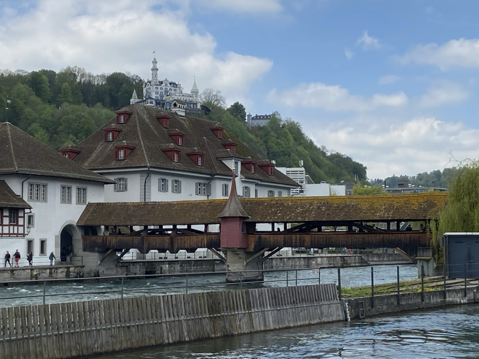 Lucerne's historic wooden Spreuer Bridge crossing the Reuss River, with the picturesque Chateau Gütsch perched on a lush green hill in the background. 