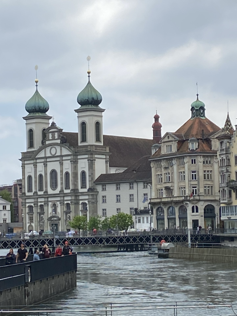 The Jesuit Church in Lucerne, a stunning Baroque-style building situated along the Reuss River. The church’s white facade is adorned with ornate details, twin green onion-shaped domes, and a bell tower. 