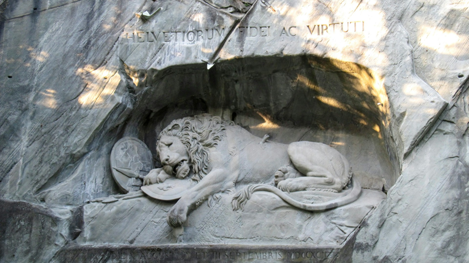 The Lion Monument in Lucerne, a large stone sculpture of a wounded lion carved into a sandstone rock face. The lion lies with a pained expression, partially draped over a shield bearing a Swiss cross, symbolizing honor and mourning. 