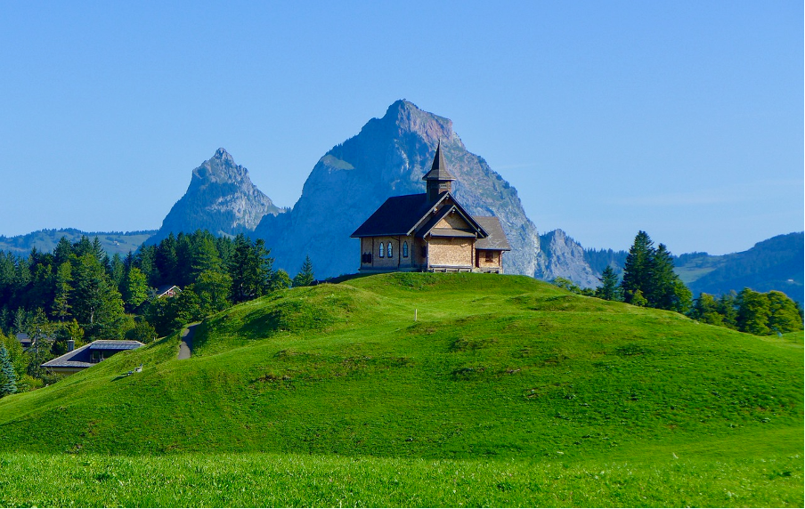 A quaint church perched atop a lush green hill in Stoos, a tranquil car-free mountain resort in Central Switzerland, surrounded by breathtaking scenery.