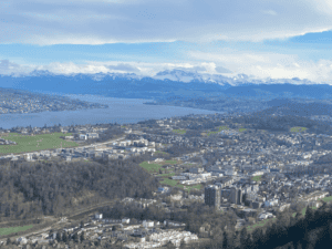 A stunning vista from Felsenegg along the Planet Trail, a scenic hike near Zurich, capturing the town of Adliswil nestled below, with Lake Zurich and the majestic, snow-covered Alps in the background.