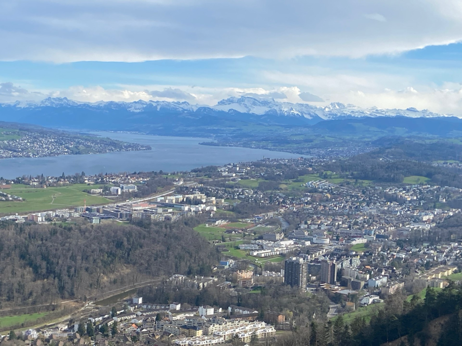 A stunning vista from Felsenegg along the Planet Trail, a scenic hike near Zurich, capturing the town of Adliswil nestled below, with Lake Zurich and the majestic, snow-covered Alps in the background.