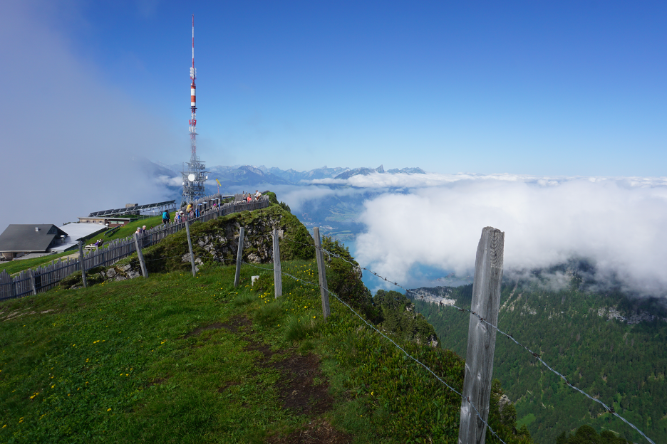 High-angle view of the summit of Mount Niederhorn, featuring a wooden fence and a tower with panoramic vistas of the surrounding mountains.