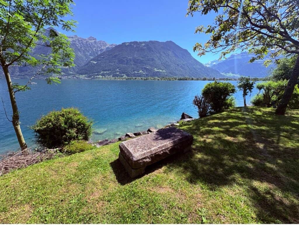 A lonely stone bench for hikers facing the blue waters of the Urnersee along the Swiss Path to Isleten.
